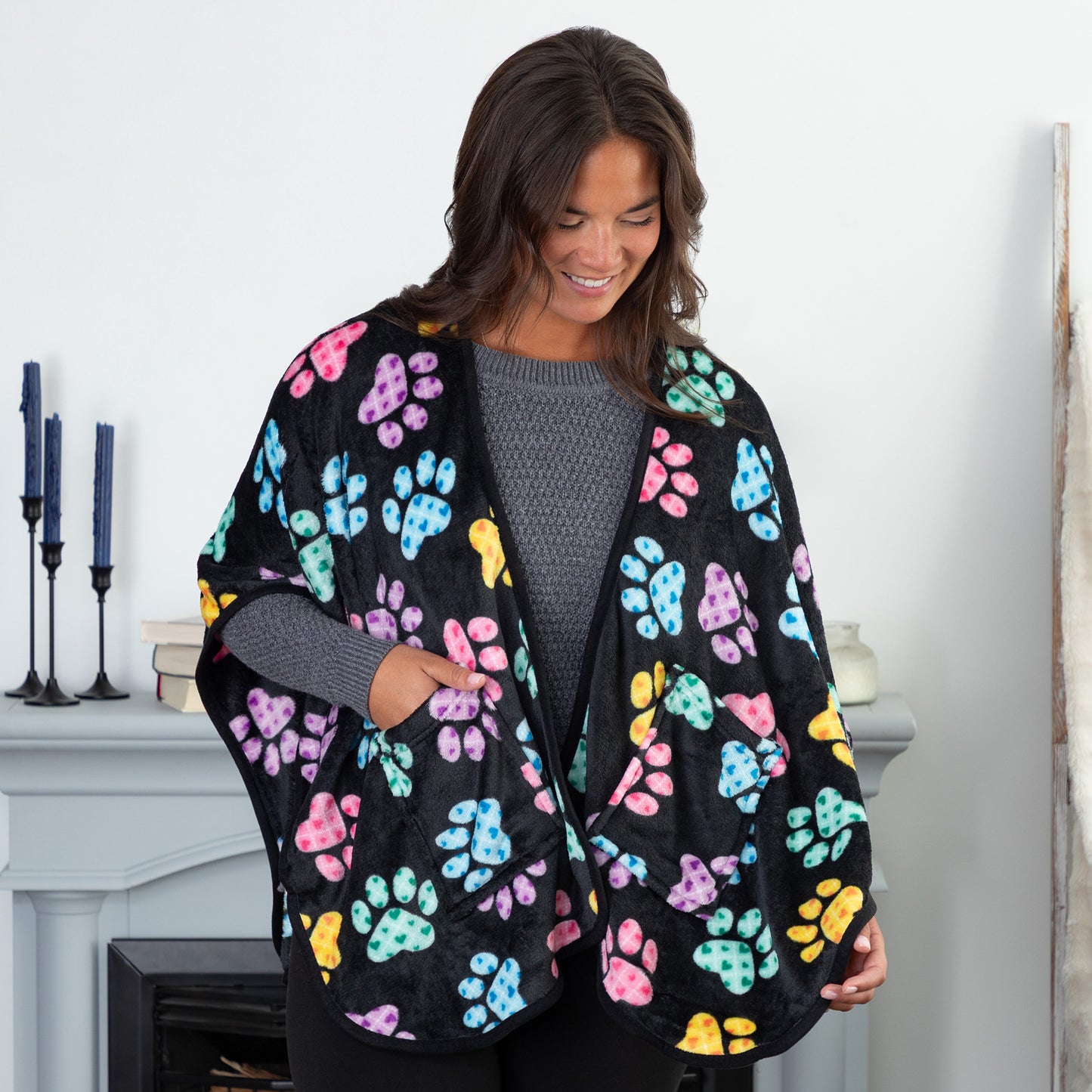 A woman standing in a cozy living room. She is wearing a black fleece, pocketed shawl, patterned with pink, blue, yellow, green, and purple argyle paw prints. 