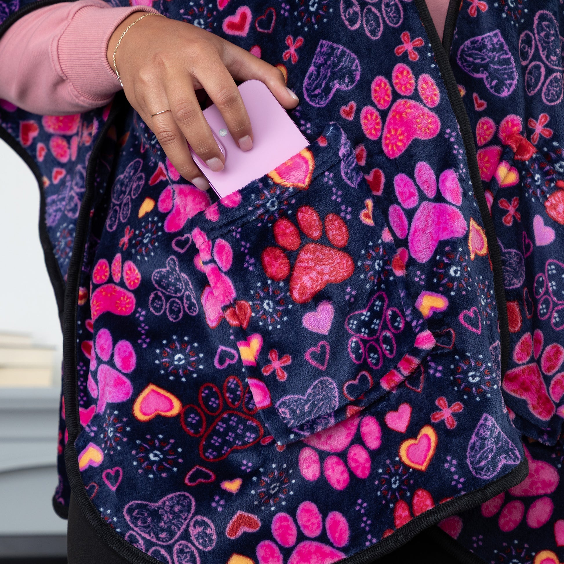 An average sized phone fitting into one of the two pockets of a deep blue based fleece blanket shawl, patterned with pink, red and purple paw prints paired with yellow, red, pink, and purple hearts. 