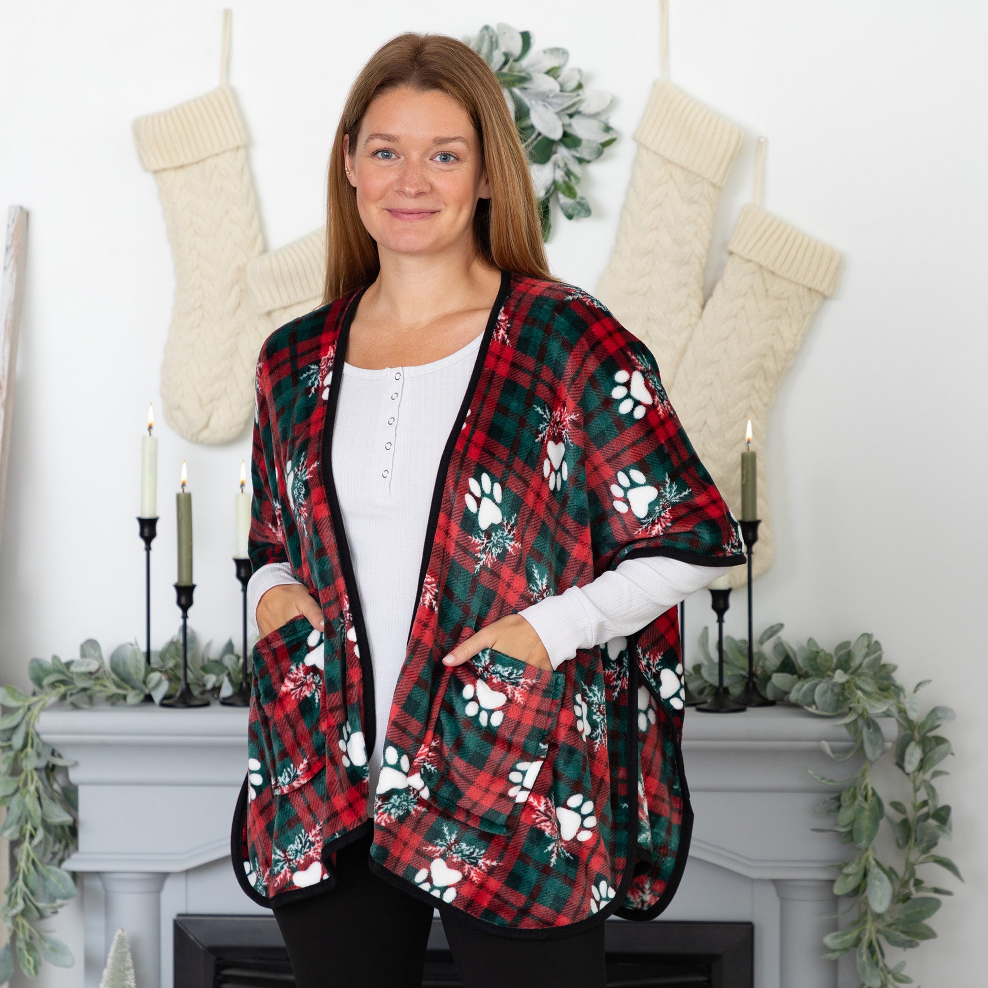 Woman in a cozy holiday setting wearing a fleece, pocketed shawl, patterned with red and green plaid overlayed with white paws and mistletoe and black piping, wrapped around her shoulders. 
