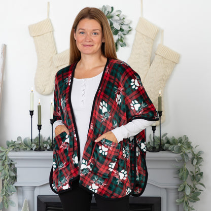 Woman in a cozy holiday setting wearing a fleece, pocketed shawl, patterned with red and green plaid overlayed with white paws and mistletoe and black piping, wrapped around her shoulders. 
