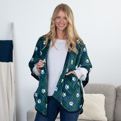 A woman stands indoors wearing a green plaid pet blanket jacket with white paw prints over a white sweater and jeans. The jacket has black trim and is shown in a living room setting with a sofa and neutral wall in the background.