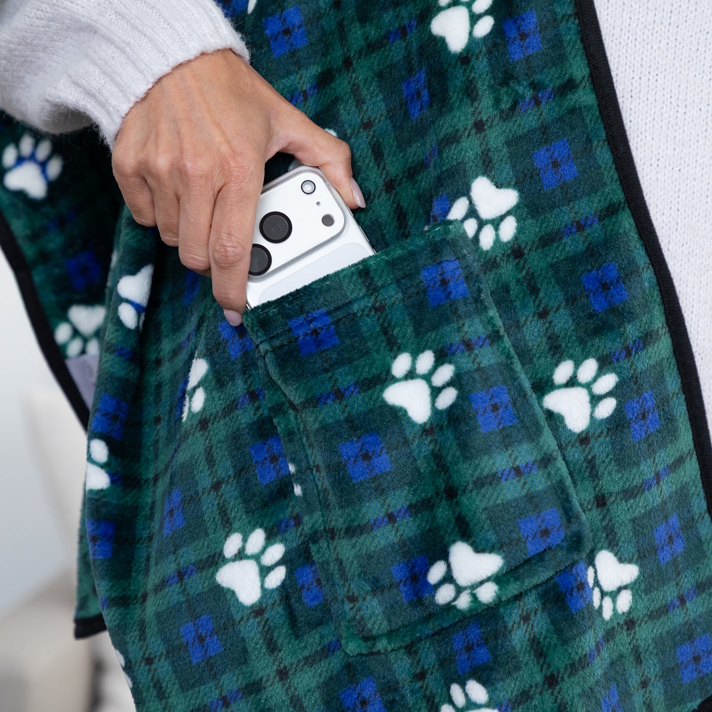 A close-up of a person wearing a green plaid blanket or throw with white paw prints and blue accents, holding a white smartphone in a front pocket.