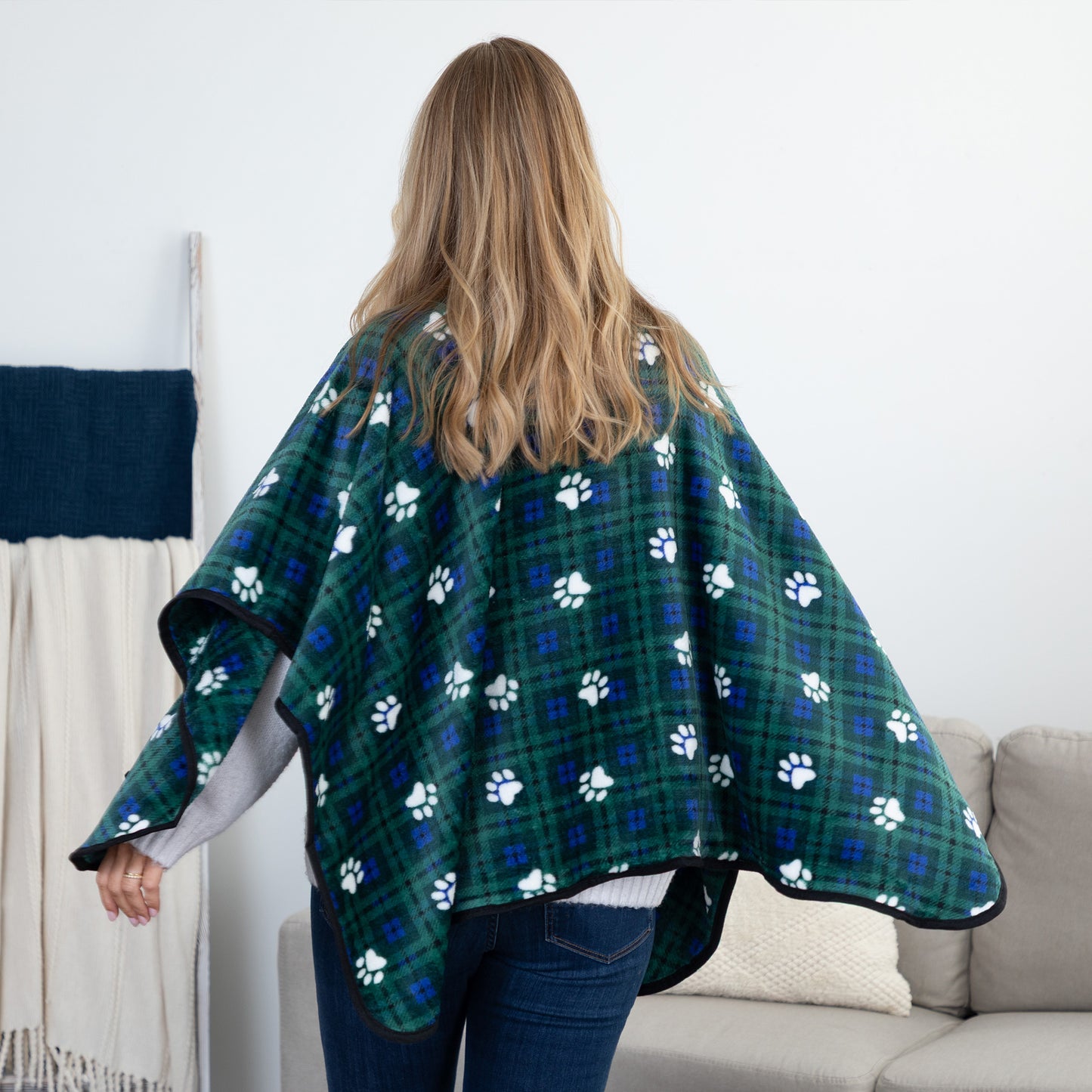 A woman viewed from behind wearing a green plaid pet blanket with white paw prints, standing in a living room next to a sofa.