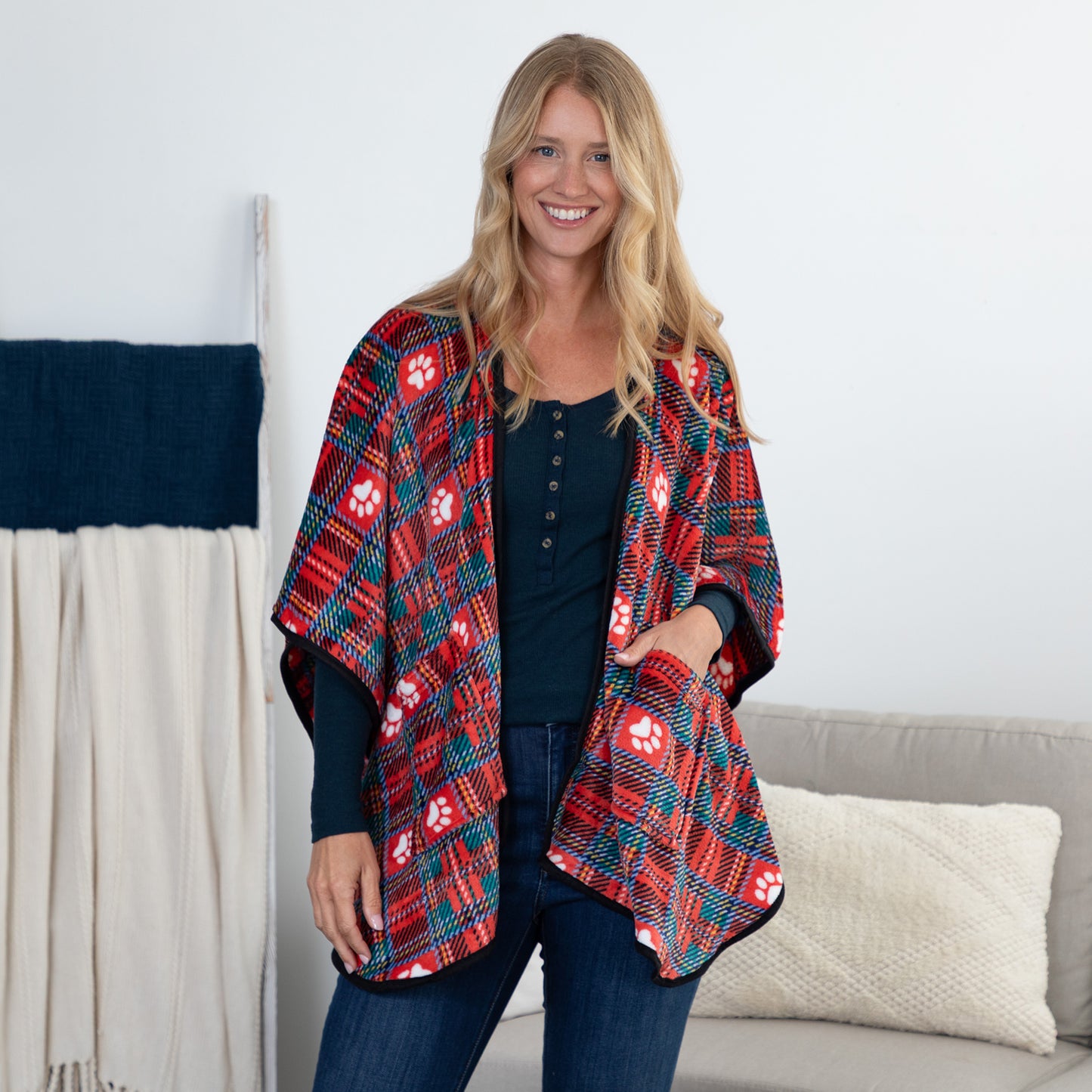 A smiling blonde woman stands indoors wearing a red and navy plaid poncho-style cardigan with white paw prints over a dark top and jeans. A light gray sofa and cream pillow are behind her, with a draped textile on a stand to the left.