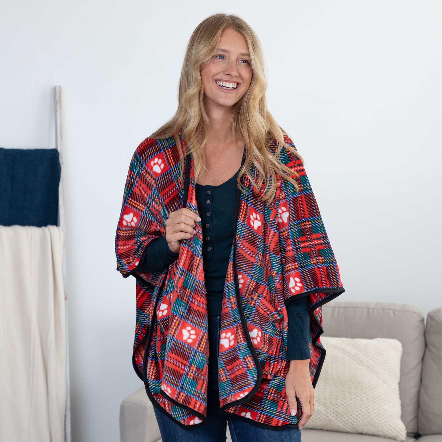 A woman stands indoors wearing a red, navy, and teal plaid poncho-style wrap with white paw print motifs over a black top. A sofa and neutral wall are visible in the background.