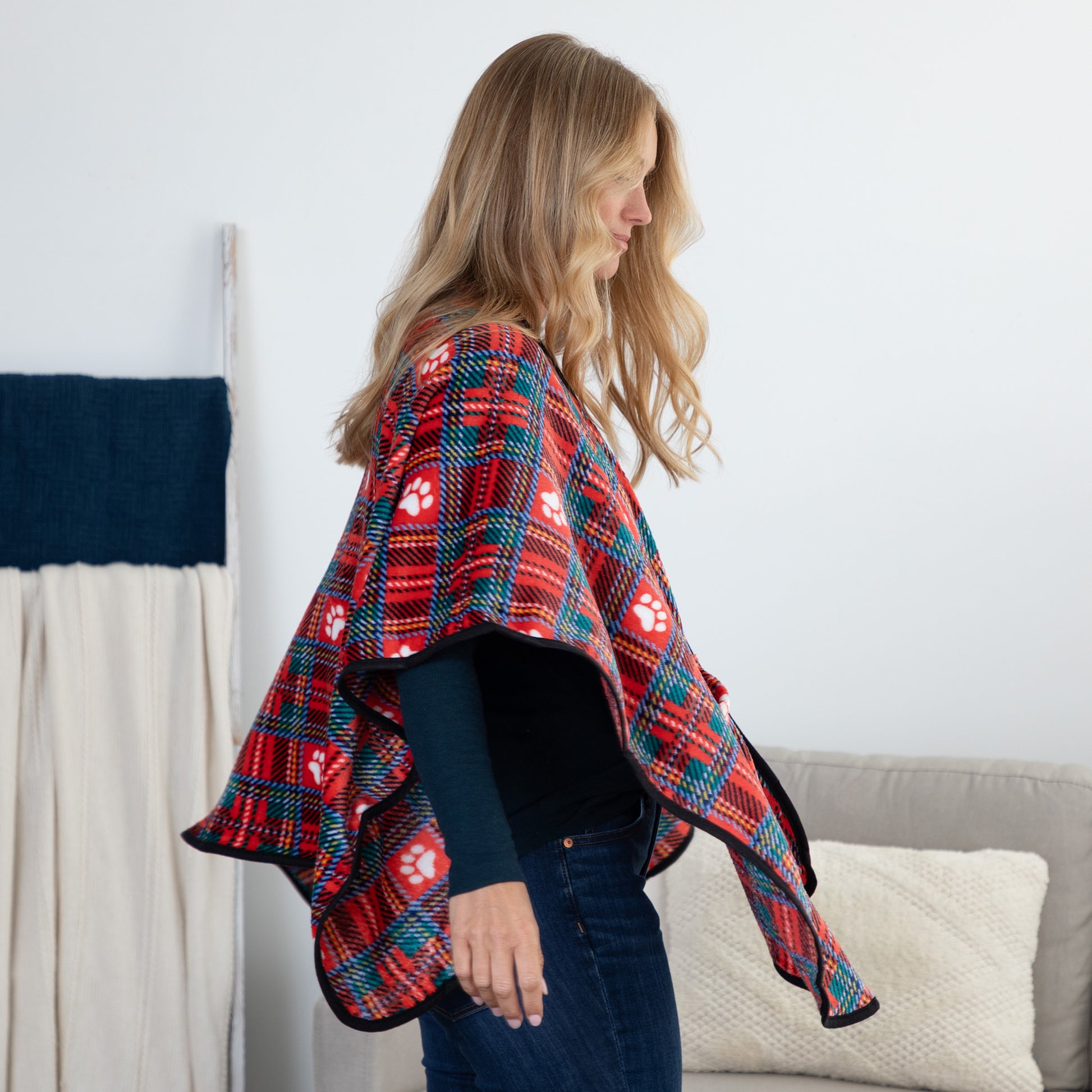 A woman wearing a red, teal, and white plaid blanket-style cape with paw print patterns, standing in a living room setting.