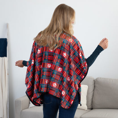 A woman with blonde hair is shown from behind wearing a red and teal plaid poncho with white paw prints, standing in a living room in front of a gray sofa.