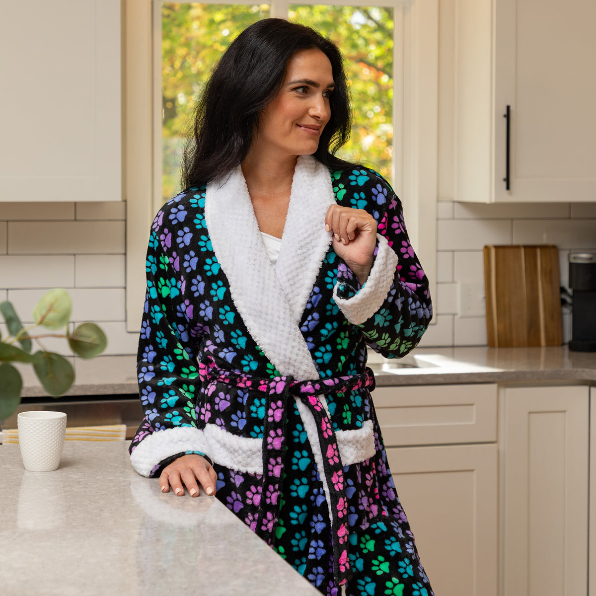 A woman wearing a plush bathrobe with a colorful paw print pattern and white sherpa trim, standing in a kitchen setting.