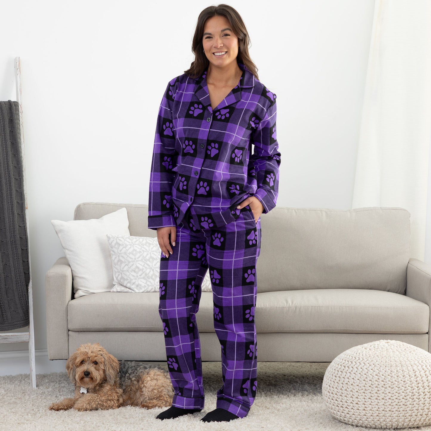 A woman standing next to her pet dog in cozy living room setting. She is wearing a matching pajama set of purple, plaid, button-up, long-sleeve top patterned with paw prints and pants of the same color and pattern. Her hand is in the side pocket of the pants. 