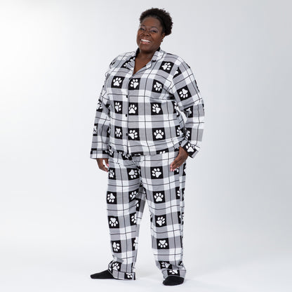 A woman wearing a matching pajama set of white and black, plaid, button-up, long-sleeve top patterned with paw prints and pants of the same color and pattern. Hand in pocket of pajamas. Plus and extended size available. 
