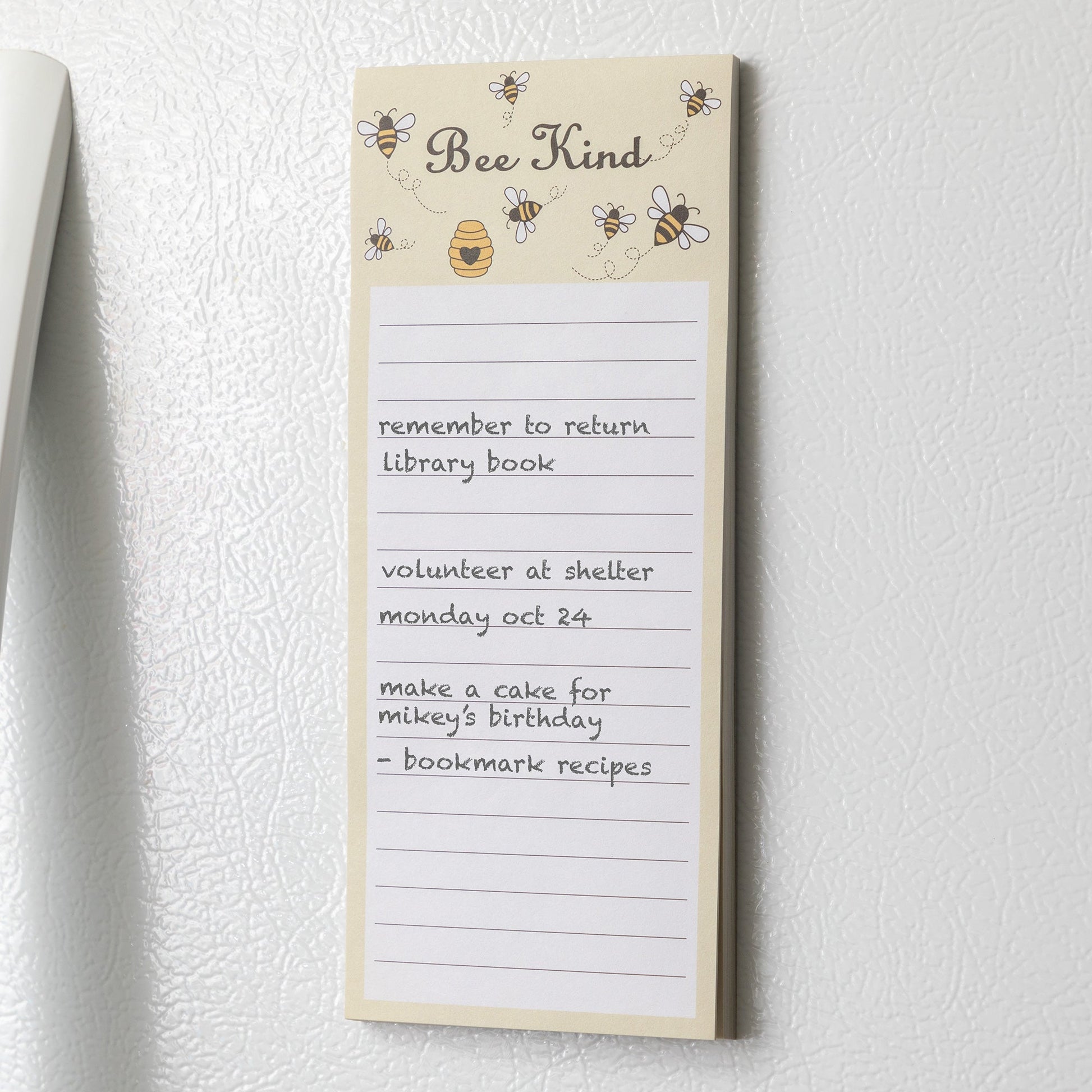 A light yellow notepad with bees and a honeycomb at the top and the phrase "Bee Kind" on a white refrigerator.