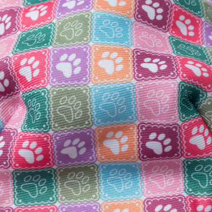 A close-up of a colorful fabric with a repeating pattern of square tiles, each containing a white paw print on a pink, purple, teal, green, and orange background.