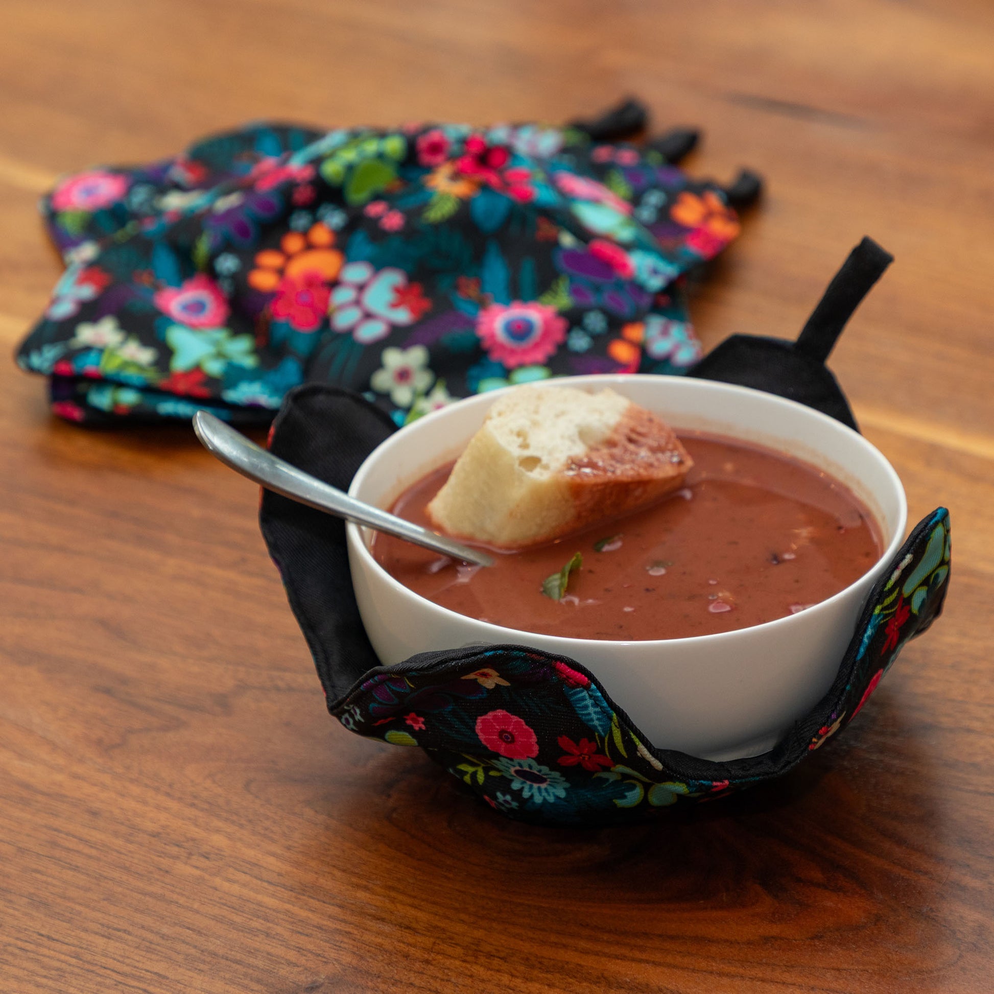 A set of four bowl cozies with various patterns, including a paw print and floral design, garden of paws, with one bowl cozy holding a bowl of hot soup and warm bread in a cozy indoor setting