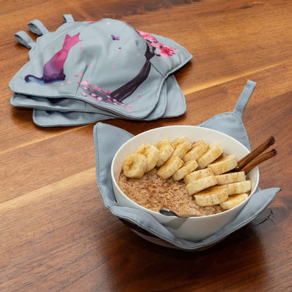A set of four bowl cozies with various patterns, including a cat and cherry blossom print and floral design, cherry blossom and cat, with one bowl cozy holding a warm bowl full of healthy oatmeal and banana