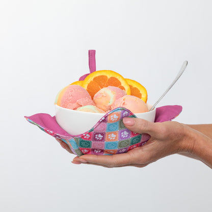 A hand holds a white bowl filled with pink and orange ice cream, garnished with orange slices, resting in a colorful patterned fabric scoop holder with a pink handle, against a plain white background.