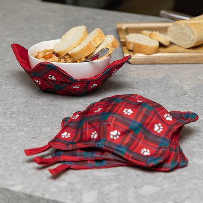Red plaid bowl cozies with white paw print pattern, shown holding a white bowl of bread on a gray countertop, with a stack of empty holders in the foreground and sliced bread on a cutting board in the background.