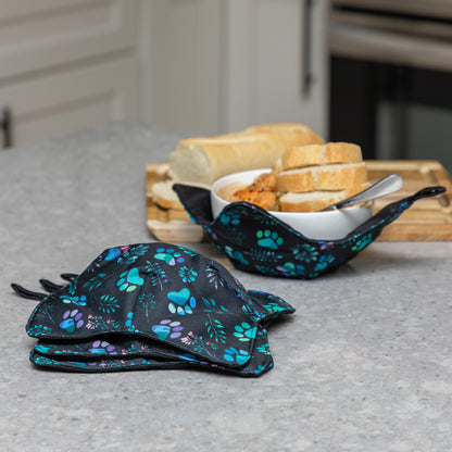 Two reusable fabric bowl cozies with a black background and blue, teal, and purple paw print pattern, shown on a kitchen counter with bread and a bowl of food in the background.