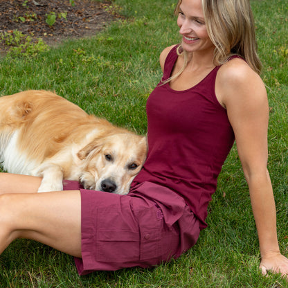 A woman sitting on green grass outdoors with a golden retriever dog beside her. She is wearing a sleeveless burgundy tank top and matching burgundy shorts.