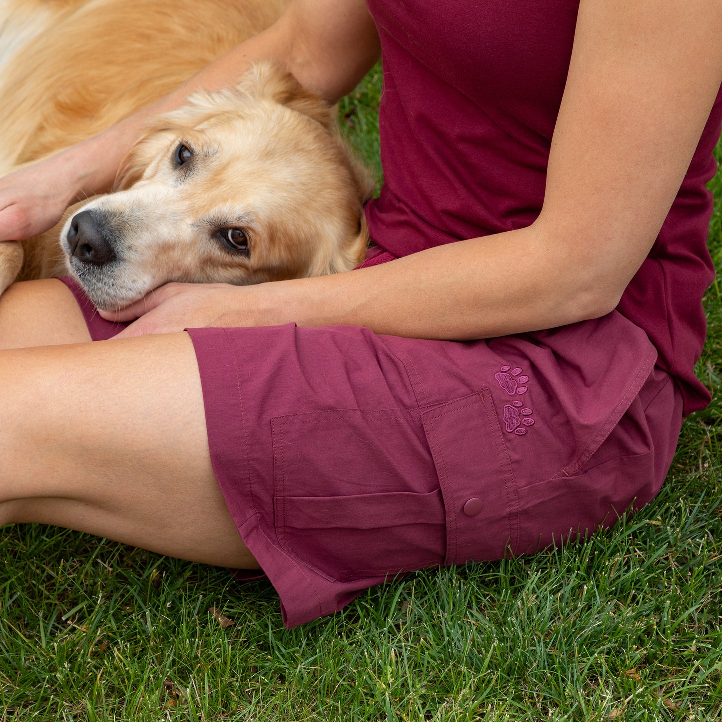 A person sitting on green grass with a golden retriever dog, wearing burgundy shorts and a matching top. The shorts feature a small embroidered logo on the pocket.