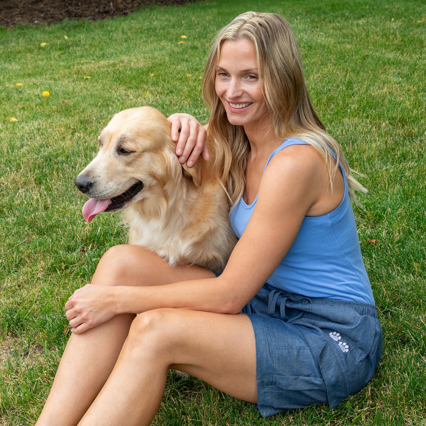 A woman sitting on green grass outdoors, petting a golden retriever dog. She is wearing a light blue tank top and denim shorts.