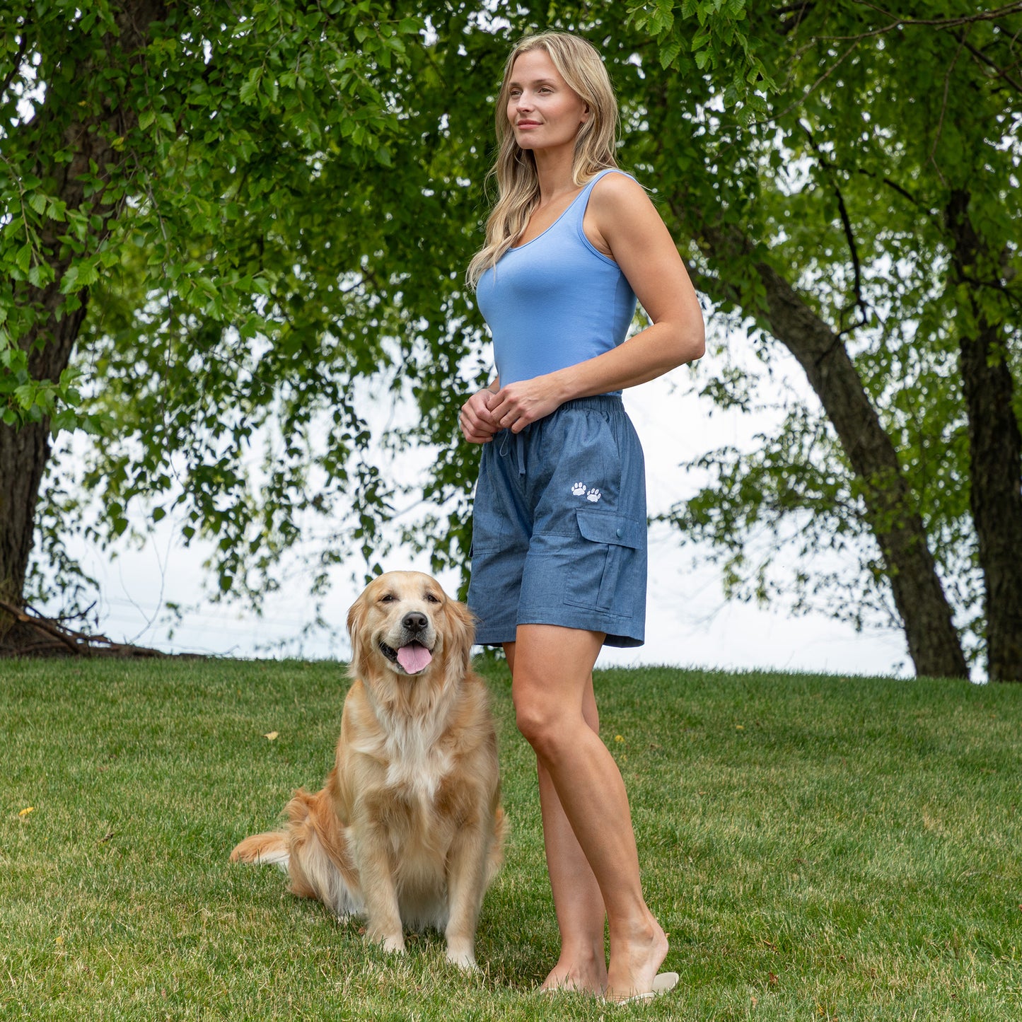 A woman wearing a light blue tank top and blue cargo shorts stands barefoot on a grassy lawn next to a golden retriever dog, with trees in the background.