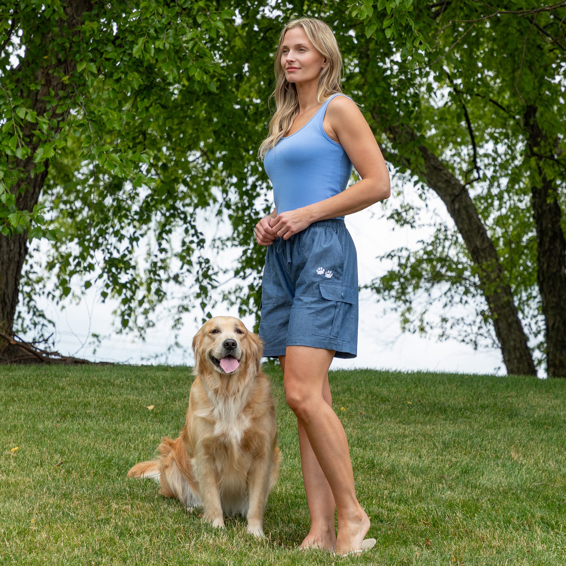 A woman wearing a light blue tank top and blue cargo shorts stands barefoot on a grassy lawn next to a golden retriever dog, with trees in the background.