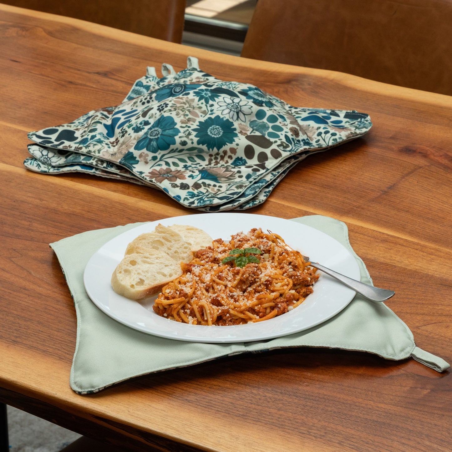 Plate of spaghetti with a bread roll on a wooden table with a paw printed stack of cozies nearby, and the plate is resting on a  plate cozy.