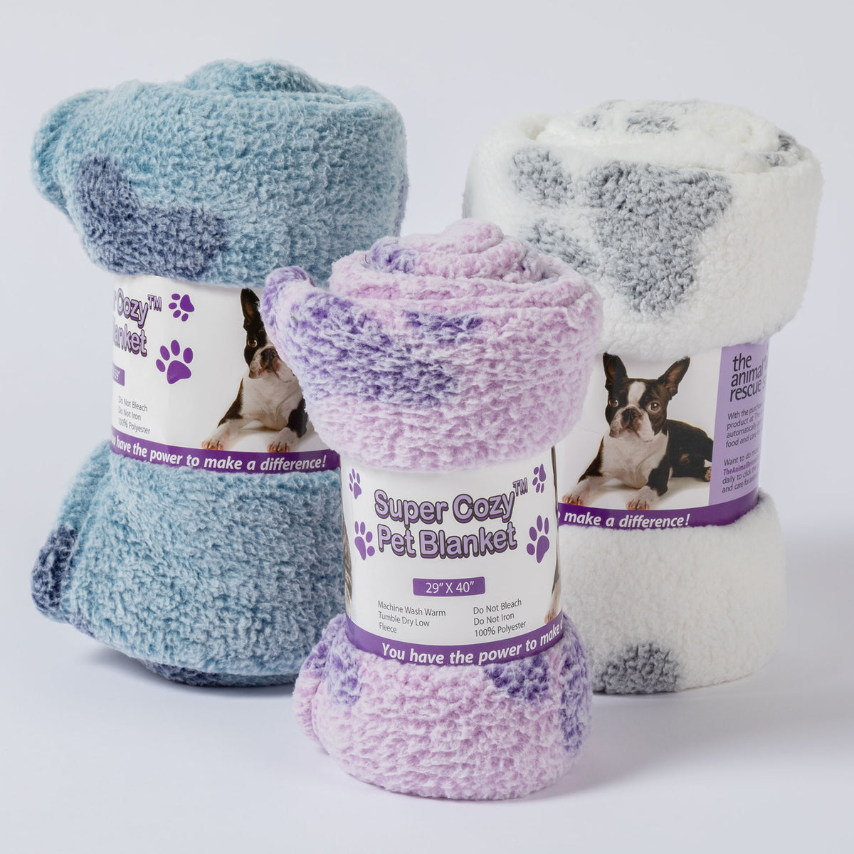 Three rolled plush pet blankets with paper labels are arranged together on a light gray studio background. The front blanket is lavender and purple, the left blanket is light blue and teal, and the right blanket is white and gray.