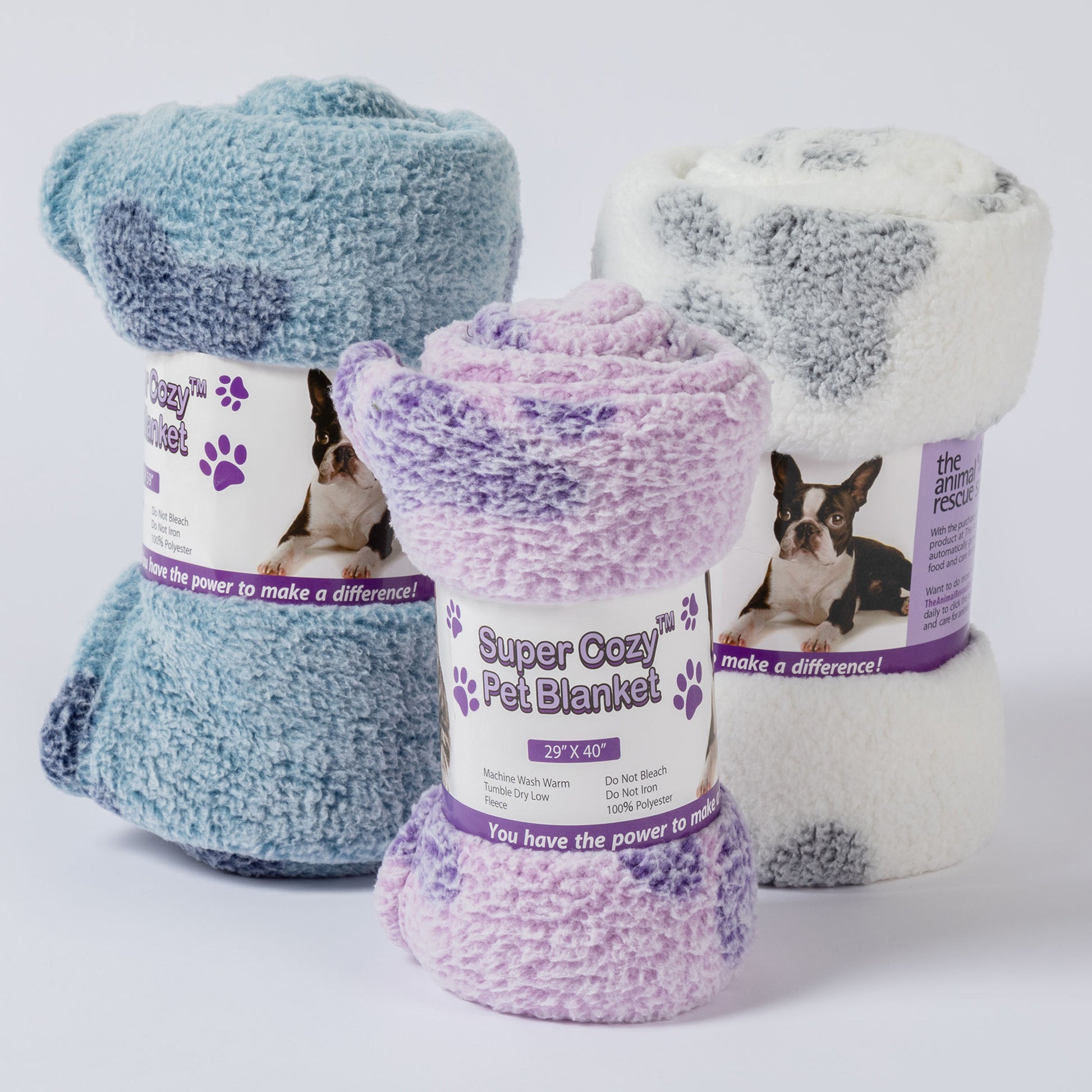 Three rolled plush pet blankets with paper labels are arranged together on a light gray studio background. The front blanket is lavender and purple, the left blanket is light blue and teal, and the right blanket is white and gray.