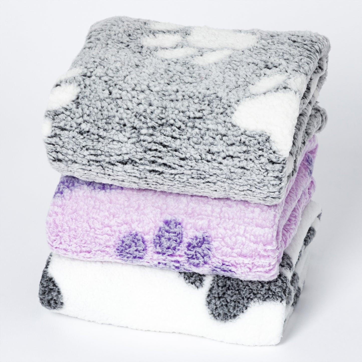Three folded plush blankets stacked on a white background: a gray blanket with white spots on top, a lavender-purple blanket with darker purple spots in the middle, and a white blanket with dark gray spots on the bottom.
