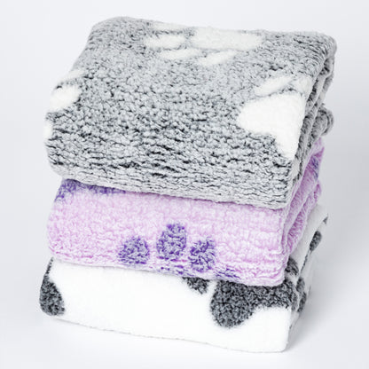 Three folded plush blankets stacked on a white background: a gray blanket with white spots on top, a lavender-purple blanket with darker purple spots in the middle, and a white blanket with dark gray spots on the bottom.