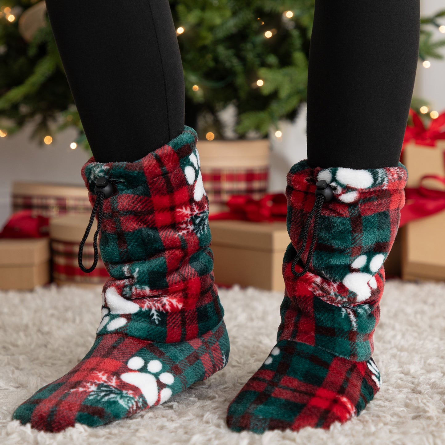 Red and green plaid slipper booties with paw prints worn indoors, Christmas decorations in the background.