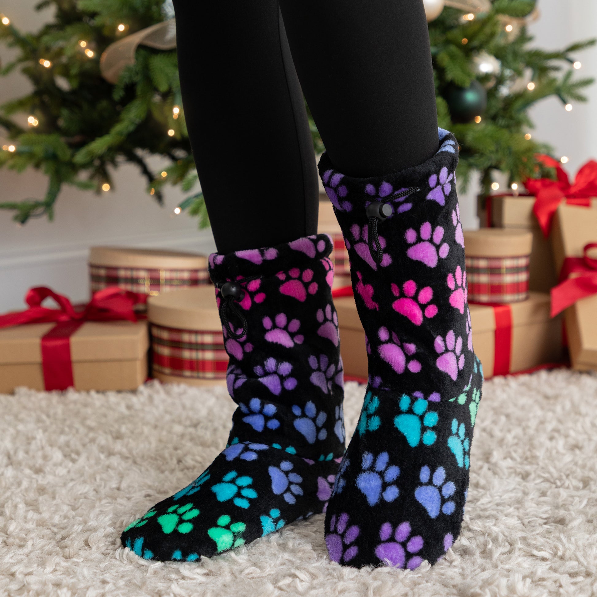 Black fleece slipper booties with colorful paw prints worn by a person in front of a decorated Christmas tree.