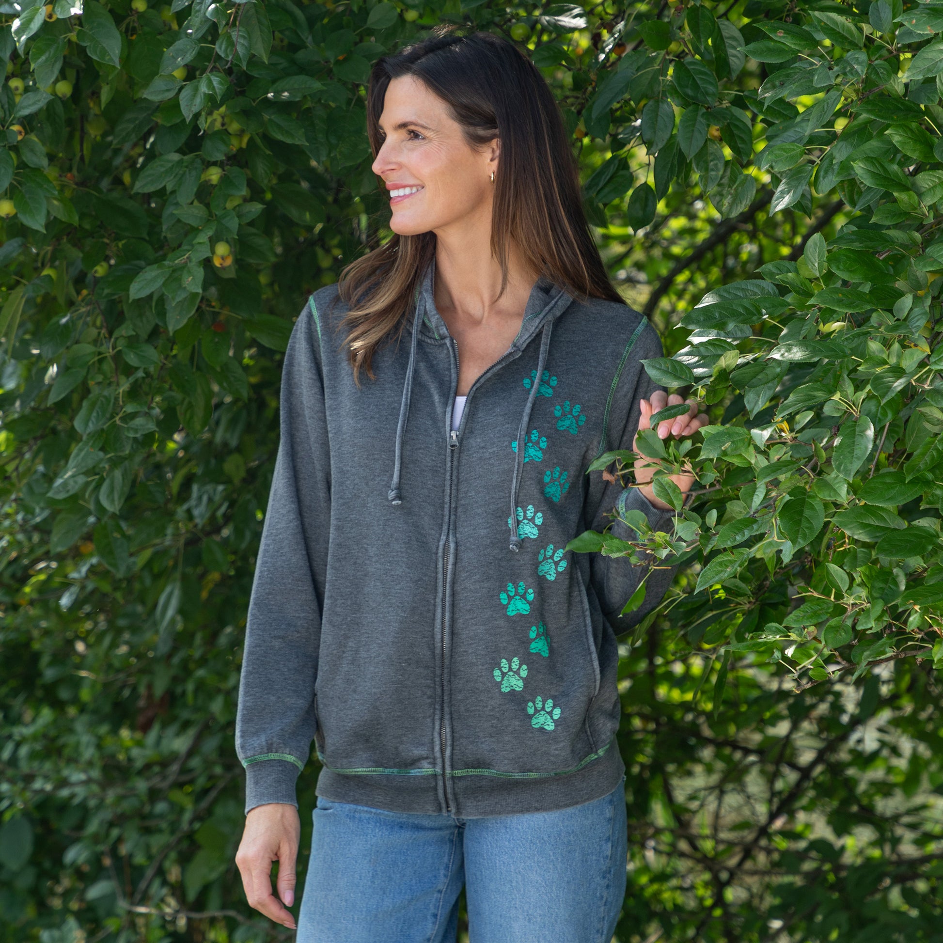 A woman wearing a dark gray zip-up hoodie with green paw print graphics on the front, standing outdoors in front of leafy green trees.