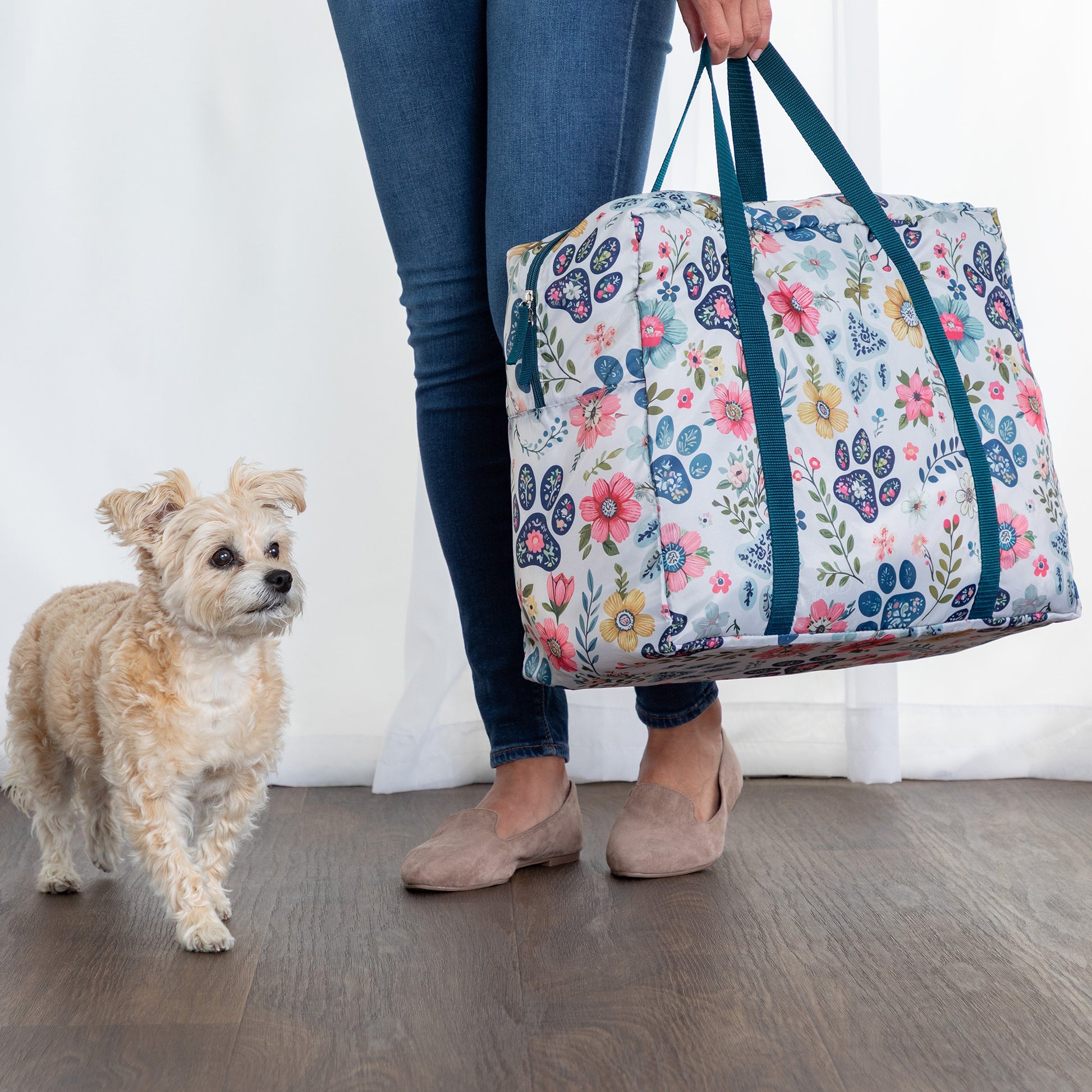 Person holding a floral-patterned duffel bag next to a small dog indoors.