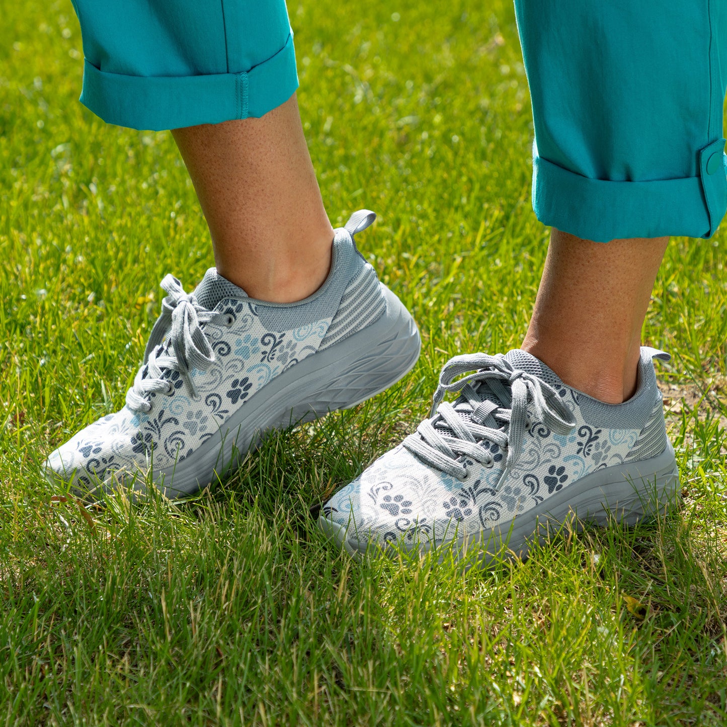 A close-up of a person wearing light gray and white patterned sneakers with teal cuffed pants, standing on a green grass lawn.