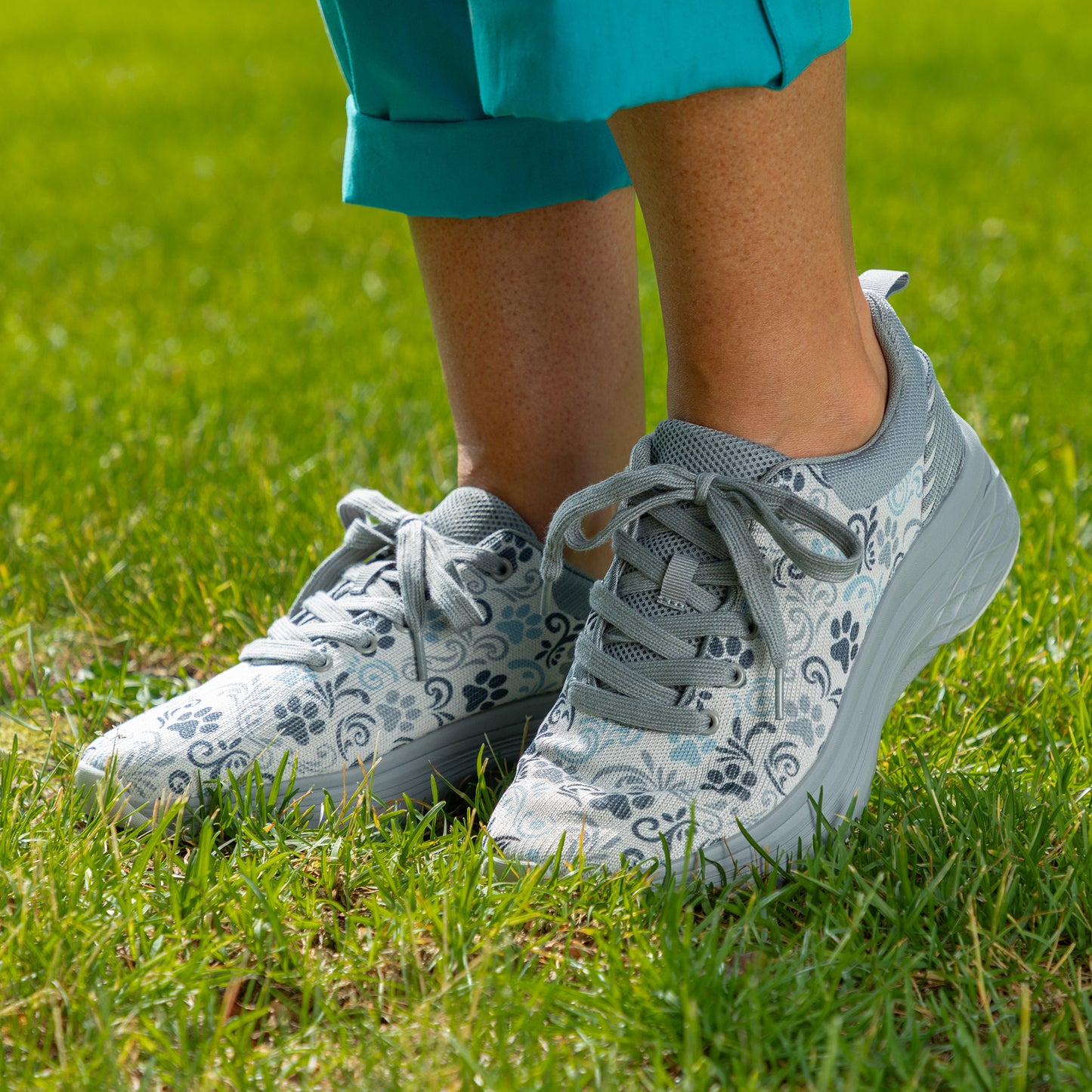 A close-up of a person wearing light gray and white floral-patterned sneakers with gray laces, standing on green grass. The person is also wearing teal cropped pants.