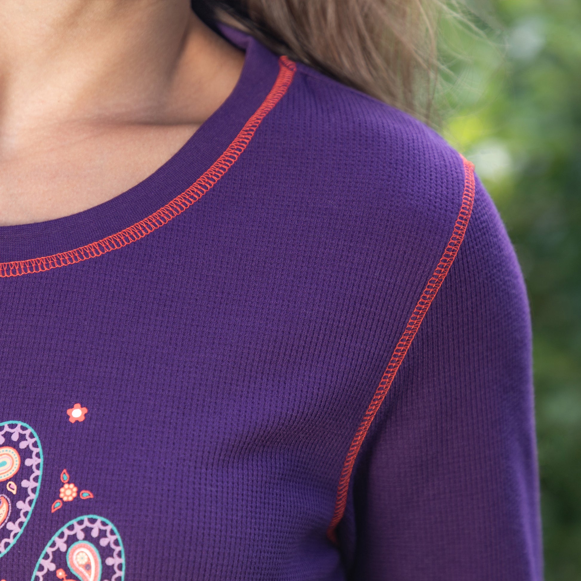 Close-up of pink contrast stitching on a purple thermal long sleeve top, with trees in the background.