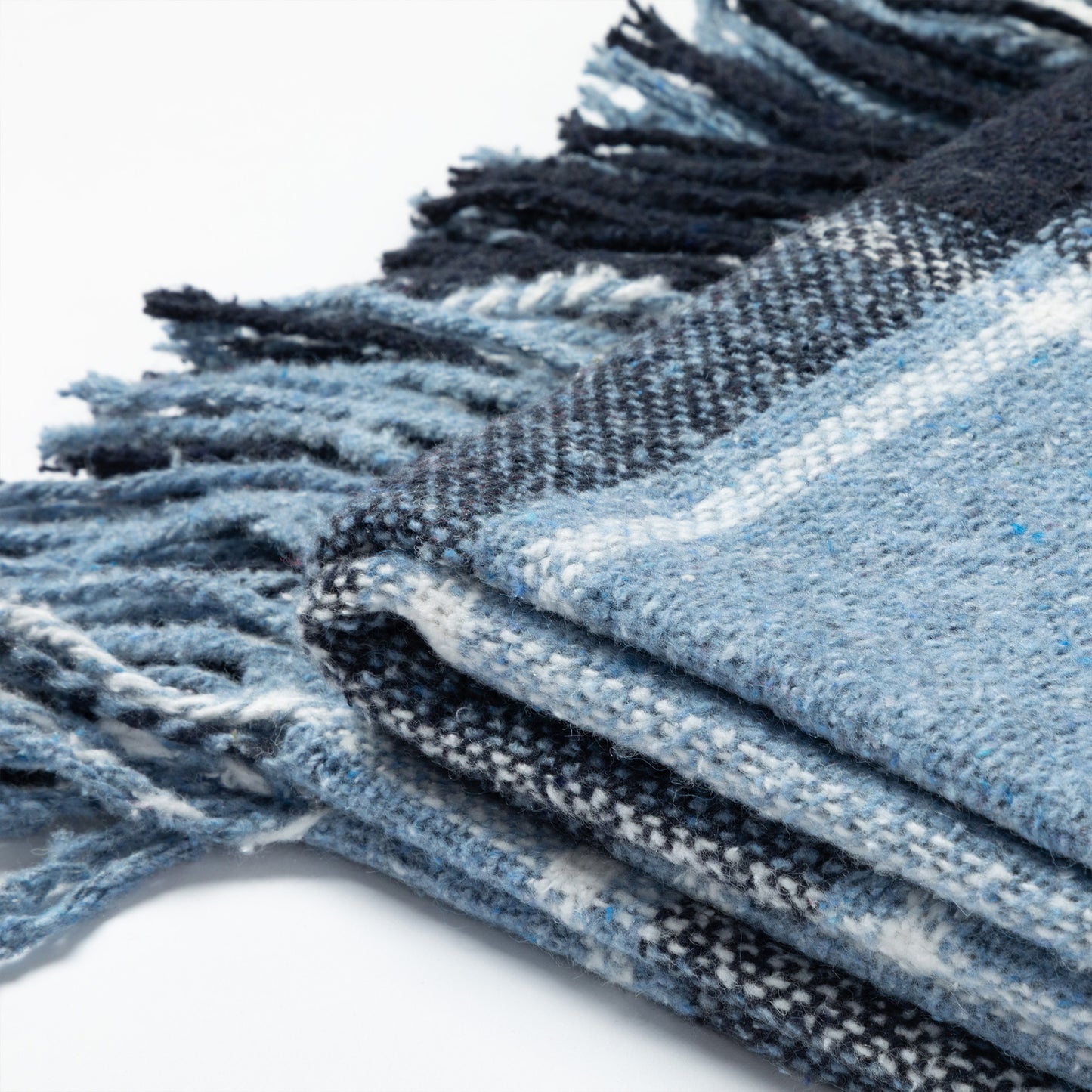 A close-up of a folded woven blanket or throw with fringed edges, showing a plaid pattern in light blue, navy, white, and gray on a white background.