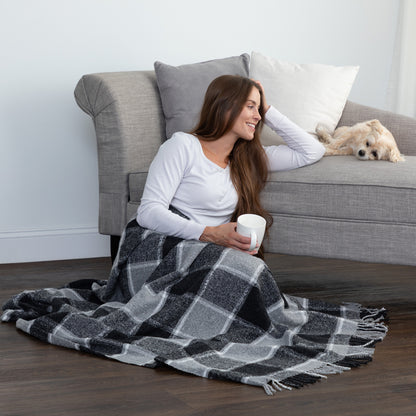 A woman sits on a dark wood floor in front of a gray sofa, wrapped in a black, gray, and white plaid blanket and holding a white mug. A small light-colored dog rests on the sofa behind her.