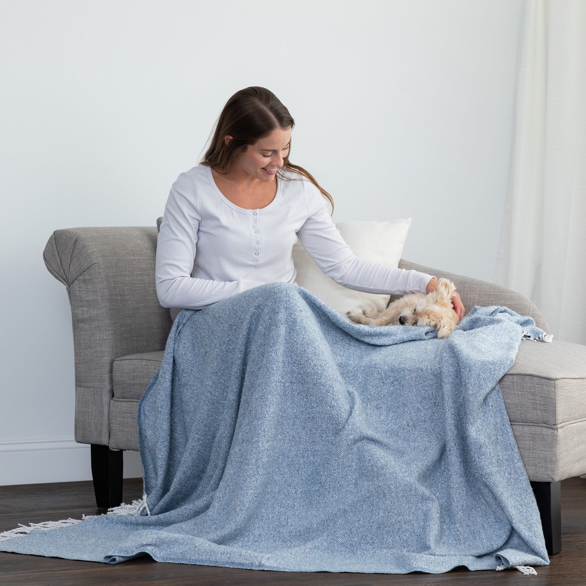 A woman sits on a gray armchair in a bright room, wrapped in a light blue textured blanket while holding a small white dog.