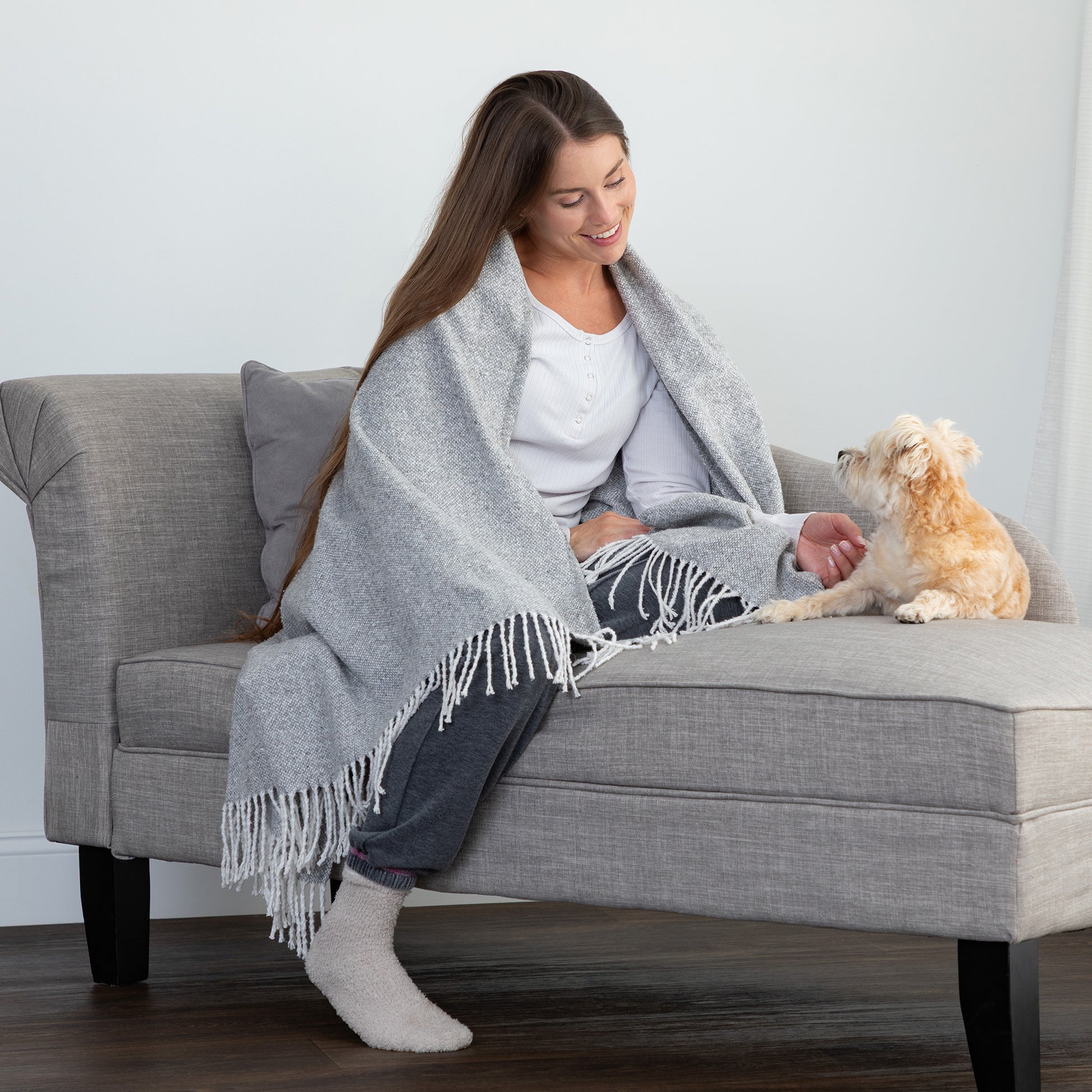 A woman sits on a gray upholstered bench or chaise wrapped in a light gray fringed throw blanket, with a small cream-colored dog beside her. The room has a light gray wall and dark wood flooring.