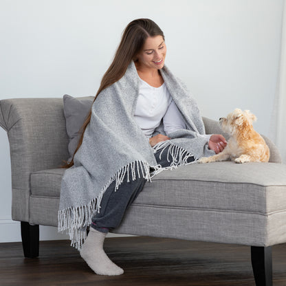 A woman sits on a gray upholstered bench or chaise wrapped in a light gray fringed throw blanket, with a small cream-colored dog beside her. The room has a light gray wall and dark wood flooring.