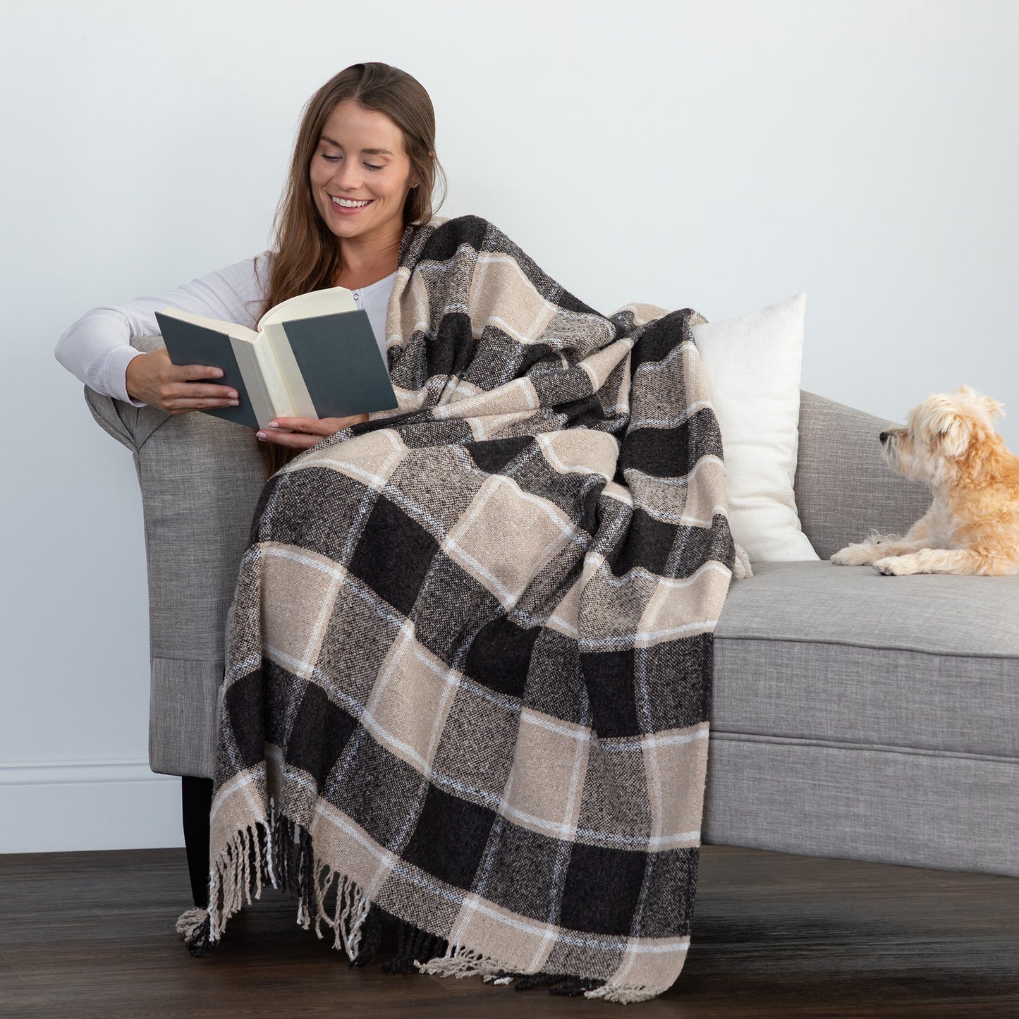 A woman sits on a gray sofa reading a book while wrapped in a large black, beige, and white plaid throw blanket, with a small light-colored dog resting on the sofa beside her.