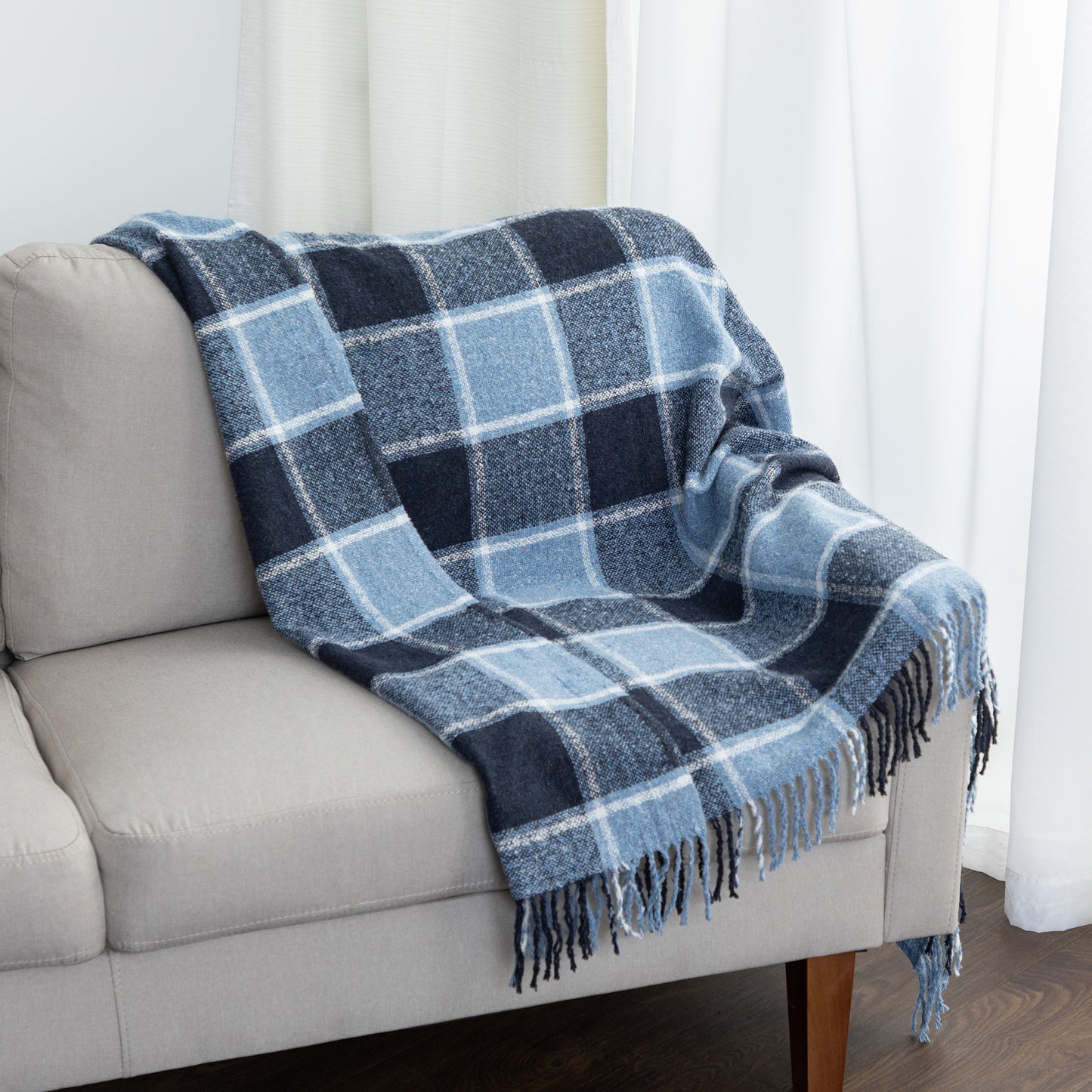 A blue and navy plaid throw blanket with fringe is draped over a light gray sofa in a bright room with white curtains.