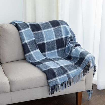 A blue and navy plaid throw blanket with fringe is draped over a light gray sofa in a bright room with white curtains.