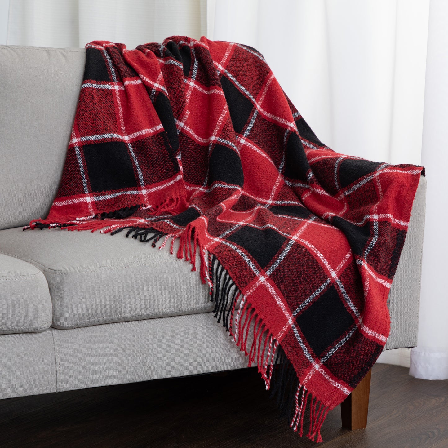 A red and black plaid throw blanket with white grid lines and fringed edges draped over a light gray sofa in a living room setting.