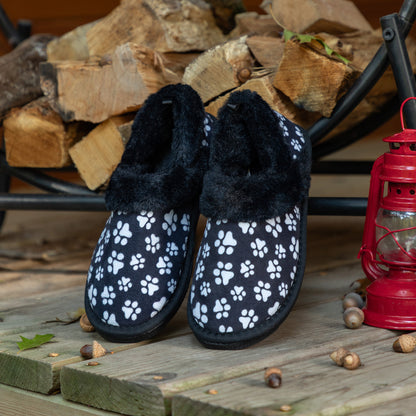 Black slippers with white paw prints on a wooden surface with firewood and a lantern in the background. Lined with black faux fur.