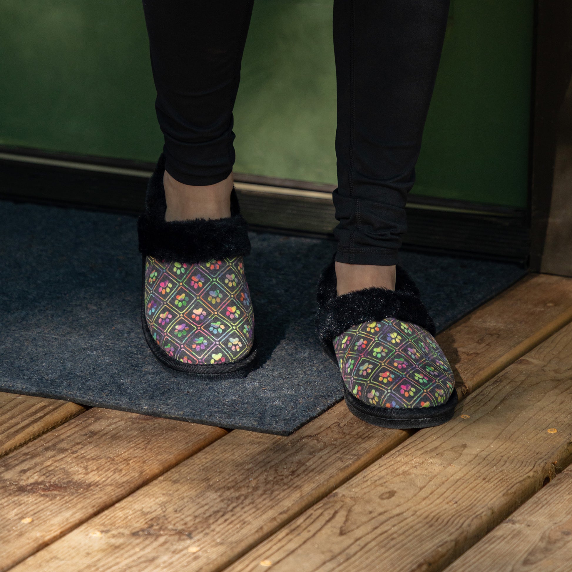 Person wearing black faux fur lined paw printed slipper shoes on wood deck.