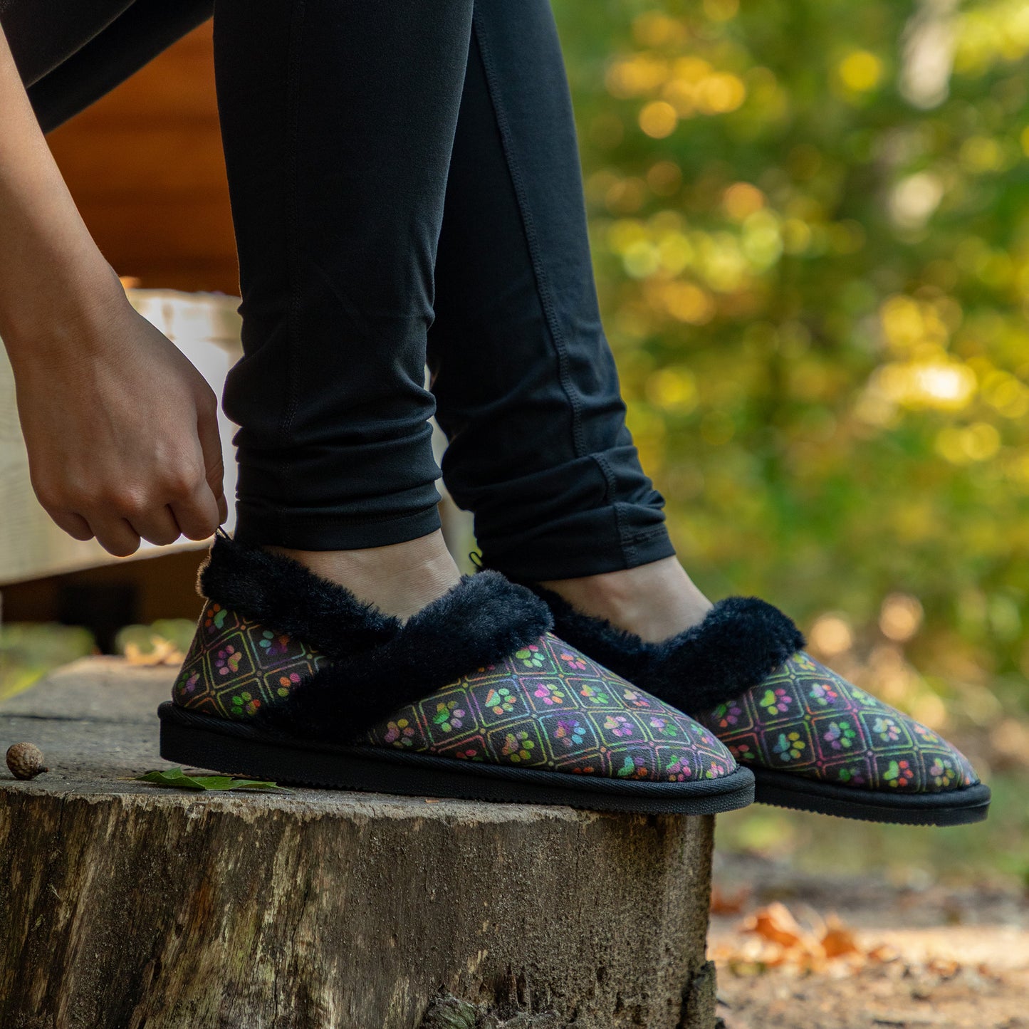 Person wearing black faux fur lined paw printed slipper shoes in outdoor wooded setting.